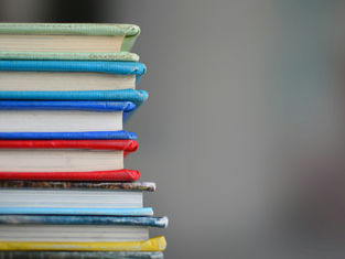 Stack of colorful books with green, blue, red, and yellow spines, against a soft, blurred gray background.