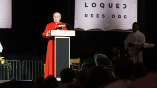 A man in red robes speaks at a podium on stage, flanked by banners and people. A sign reads "MAS DE LO QUE CREES.ORG" in the background.