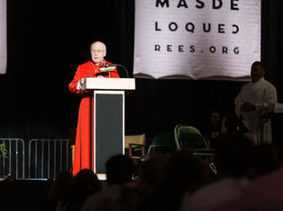 A man in red robes speaks at a podium on stage, flanked by banners and people. A sign reads "MAS DE LO QUE CREES.ORG" in the background.