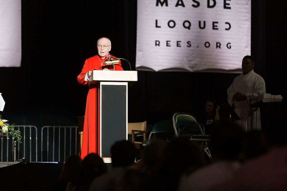 A man in red robes speaks at a podium on stage, flanked by banners and people. A sign reads "MAS DE LO QUE CREES.ORG" in the background.