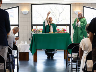 Priest lifts the Eucharist at a church altar as congregants stand and pray, with green vestments, candles, and flowers.