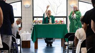 Priest lifts the Eucharist at a church altar as congregants stand and pray, with green vestments, candles, and flowers.
