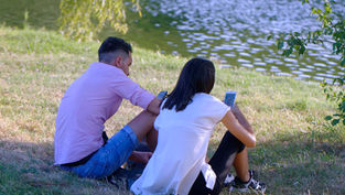 Two people sit on grass by a pond, engrossed in their phones. One wears a pink shirt and denim shorts, the other a white top and black pants.