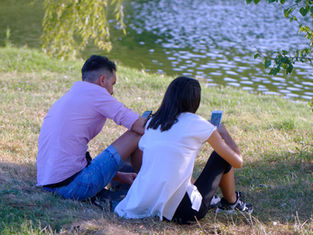 Two people sit on grass by a pond, engrossed in their phones. One wears a pink shirt and denim shorts, the other a white top and black pants.