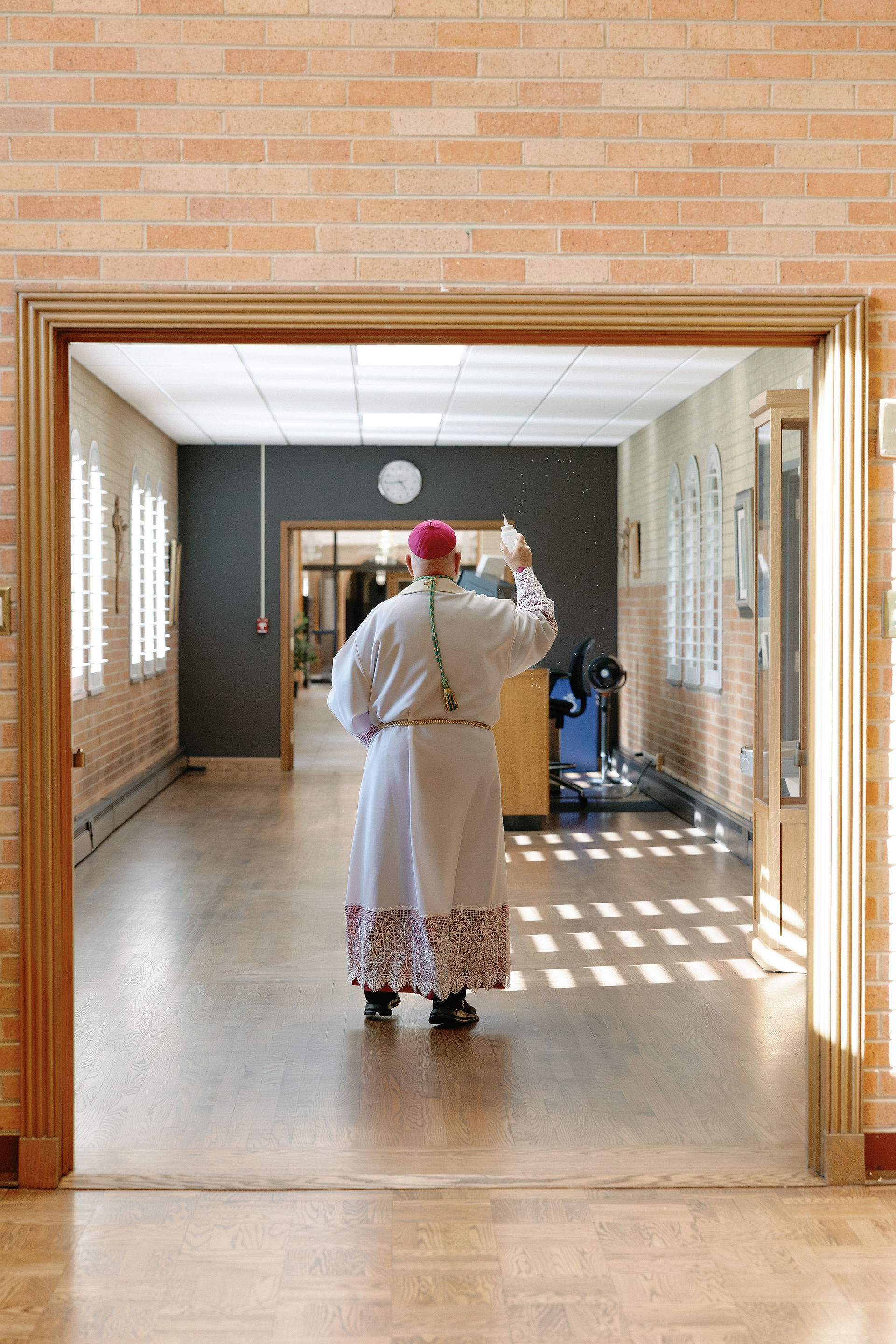 A person in clerical attire sprinkles holy water in a sunlit room with brick walls, a fan, and a clock. Quiet, reverent atmosphere.