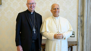 Two smiling clergymen pose indoors, one in black suit and one in white papal robes, before cream patterned wallpaper.