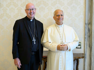 Two smiling clergymen pose indoors, one in black suit and one in white papal robes, before cream patterned wallpaper.