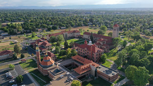 Aerial view of a campus with red-roofed buildings, green lawns, and trees. Mountains and city skyline in the background under clear skies.