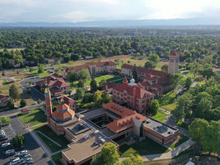 Aerial view of a campus with red-roofed buildings, green lawns, and trees. Mountains and city skyline in the background under clear skies.