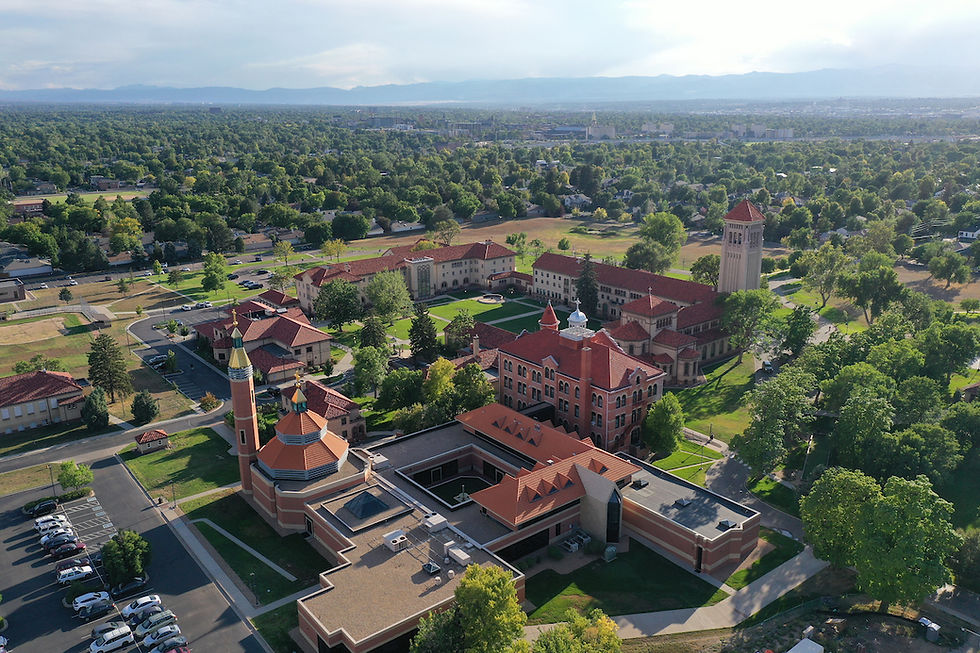 Aerial view of a campus with red-roofed buildings, green lawns, and trees. Mountains and city skyline in the background under clear skies.