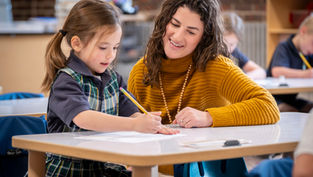 Teacher assists smiling girl writing at a classroom table. The teacher wears a mustard sweater. Background shows blurred students and books.