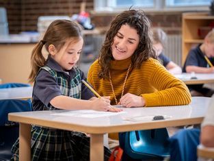 Teacher assists smiling girl writing at a classroom table. The teacher wears a mustard sweater. Background shows blurred students and books.