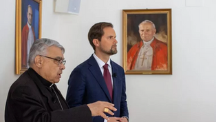 Two men in suits stand indoors near framed portraits of religious figures in red robes. One gestures while speaking, creating a solemn mood.