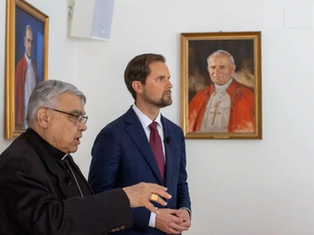 Two men in suits stand indoors near framed portraits of religious figures in red robes. One gestures while speaking, creating a solemn mood.