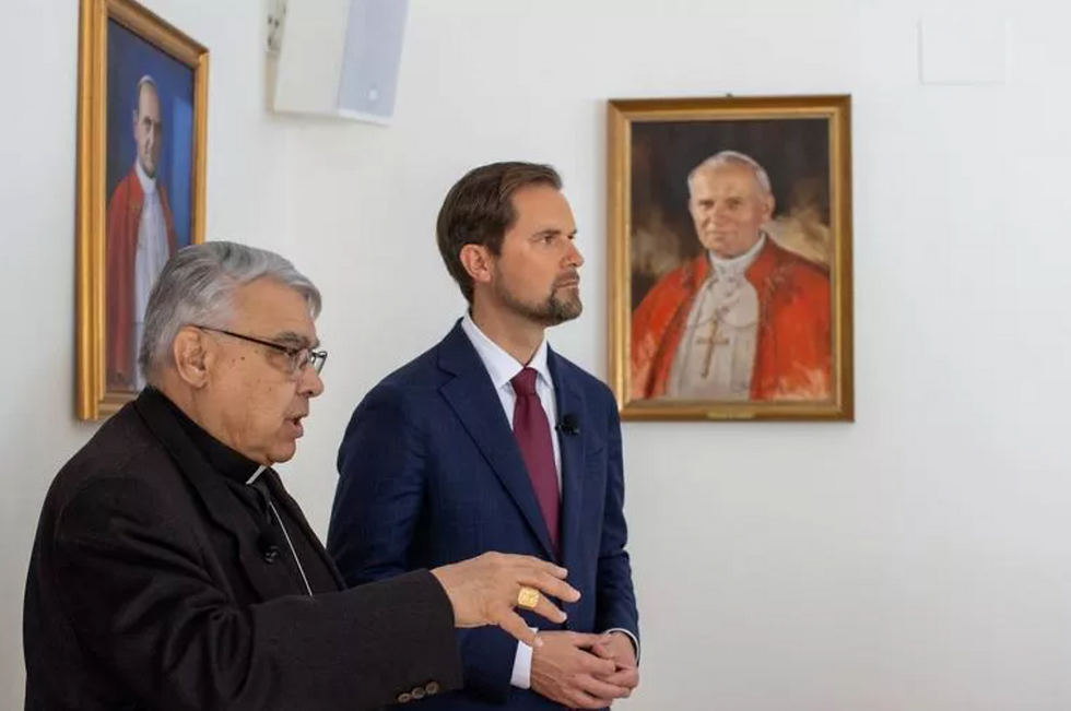 Two men in suits stand indoors near framed portraits of religious figures in red robes. One gestures while speaking, creating a solemn mood.
