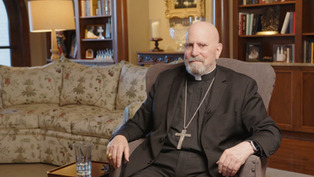 Man in black religious attire with a cross necklace sits in an armchair. Background has floral sofa, bookshelves, and a lamp. Cozy setting.
