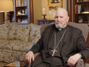 Man in black religious attire with a cross necklace sits in an armchair. Background has floral sofa, bookshelves, and a lamp. Cozy setting.