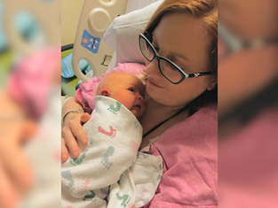 Mother in glasses cradling newborn wrapped in a patterned blanket. Hospital setting. Pink and white tones, tender and calm mood.