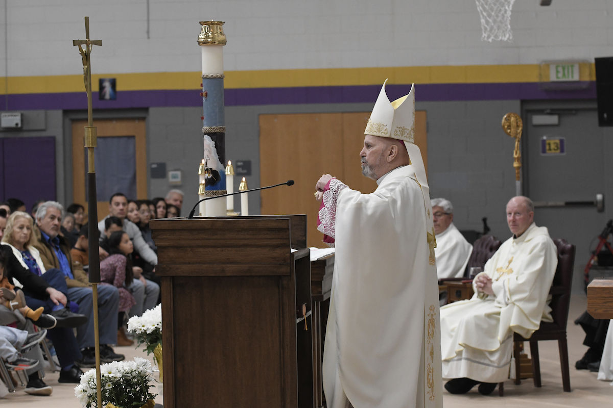 Clergy in white robes conducting a service in a gymnasium with a seated audience. Tall candle and cross visible. Solemn atmosphere.