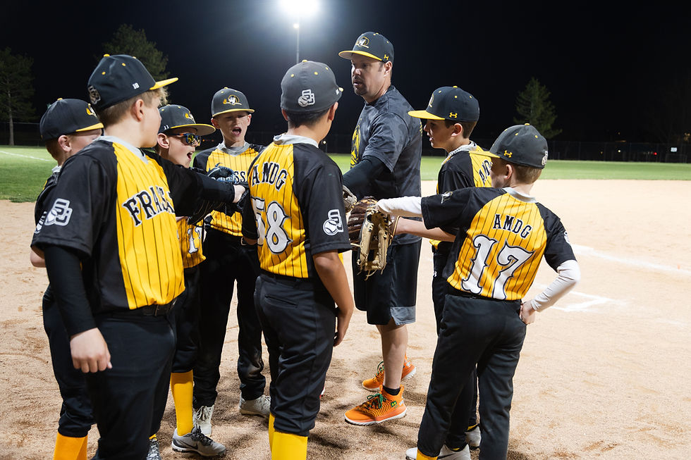 Youth baseball team in yellow jerseys huddles with coach at night on a lit field, showing teamwork and enthusiasm.