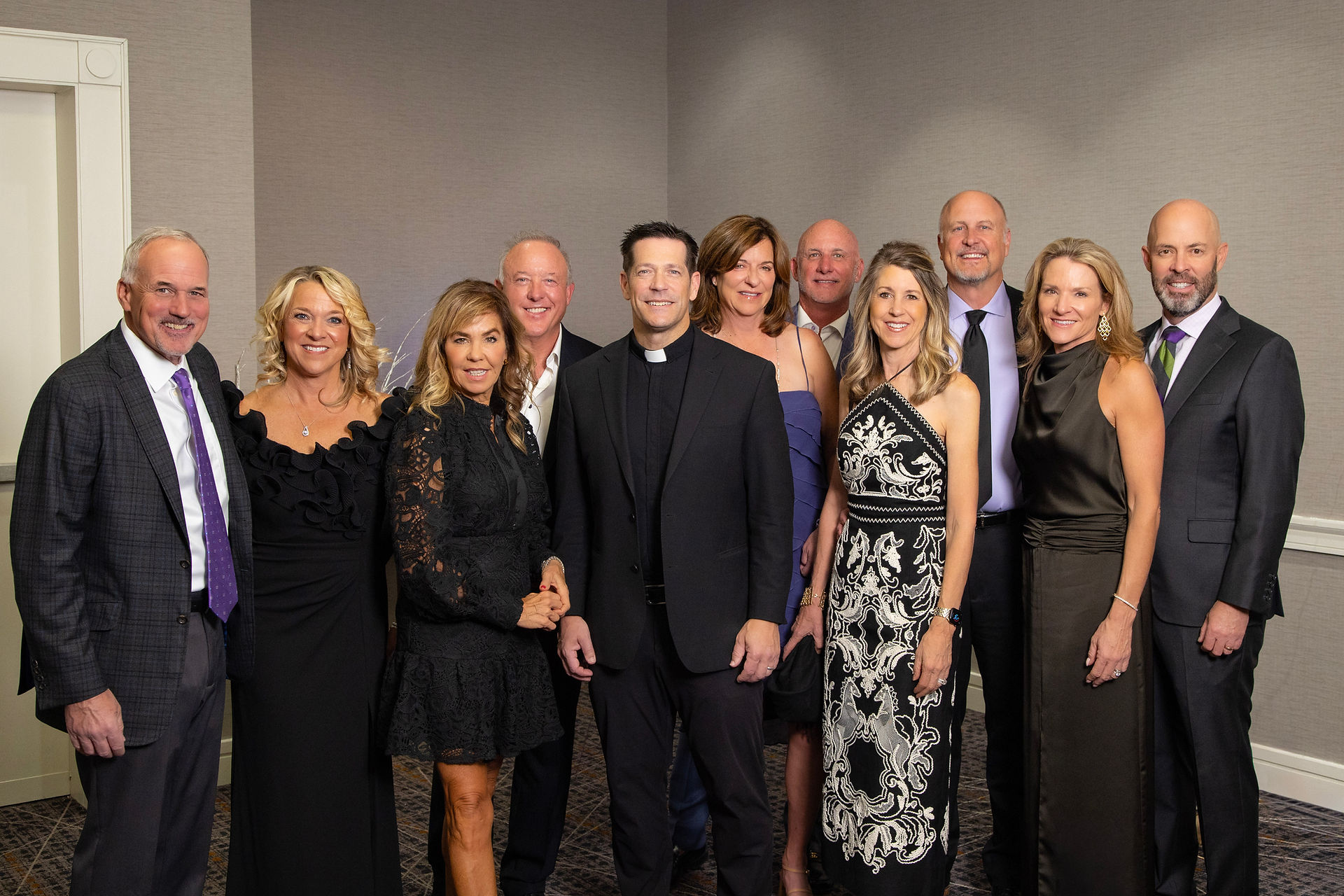A group of ten well-dressed people pose in a formal setting with neutral walls. They smile warmly, exuding a joyful and elegant atmosphere.