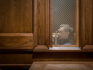 Man seen through a confessional screen in a wooden booth. The setting is dimly lit, conveying a solemn, contemplative mood.