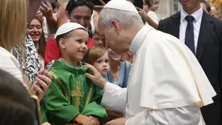 The image shows a religious figure smiling warmly at a young boy in a green robe with a gold cross, surrounded by a crowd in a church.