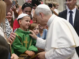 The image shows a religious figure smiling warmly at a young boy in a green robe with a gold cross, surrounded by a crowd in a church.