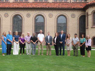 Twelve people stand in a row on a lawn before a brick building with arched windows, posing for a formal group photo.