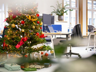 Decorated Christmas tree with lights, red and gold ornaments, and gifts underneath in an office setting with desks and chairs. Festive mood.