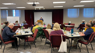 People sit at tables, listening to a nun speaking from a podium. A whiteboard reads "Welcome to the Gospel of Life Workshop."