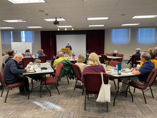 People sit at tables, listening to a nun speaking from a podium. A whiteboard reads "Welcome to the Gospel of Life Workshop."