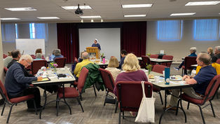 People sit at tables, listening to a nun speaking from a podium. A whiteboard reads "Welcome to the Gospel of Life Workshop."