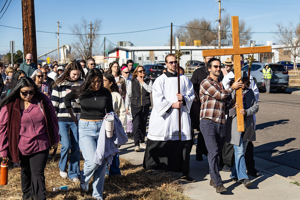 A group of people, including a person in a robe, walk down a street carrying a large wooden cross. The sky is clear and sunny.