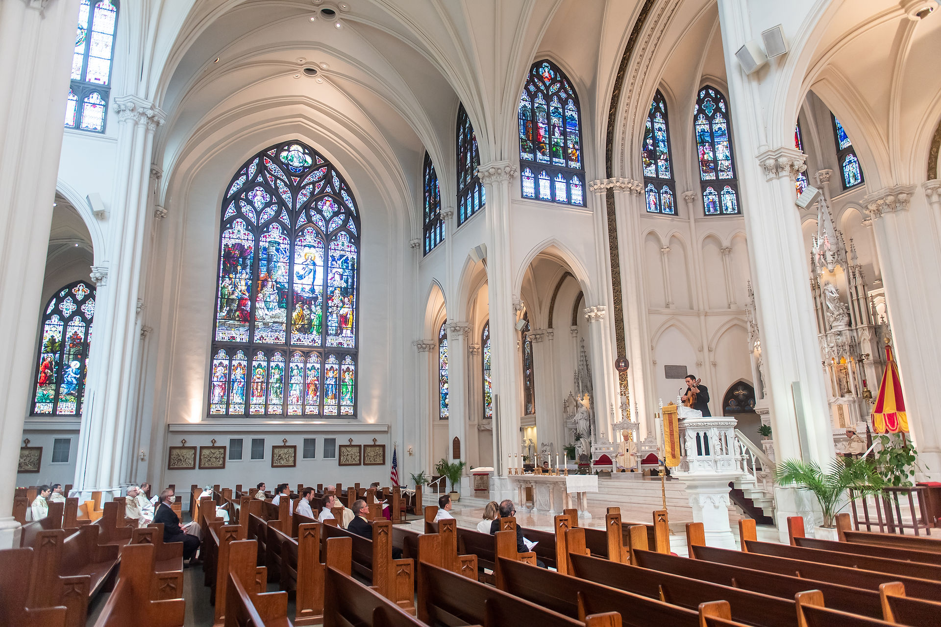 People seated in a cathedral with stained glass windows, listening to a speaker at the altar. Bright, serene atmosphere.