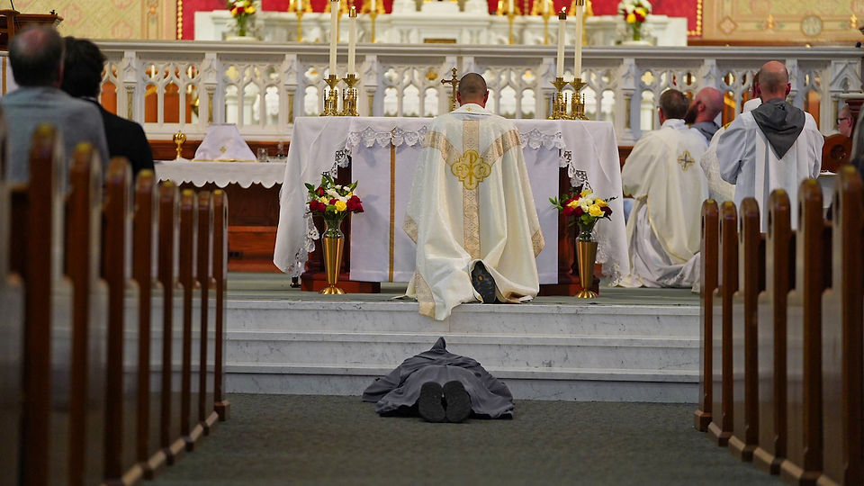 A person lies prostrate in a church aisle, facing an altar with a kneeling priest in a white robe. Candles and flowers adorn the scene.