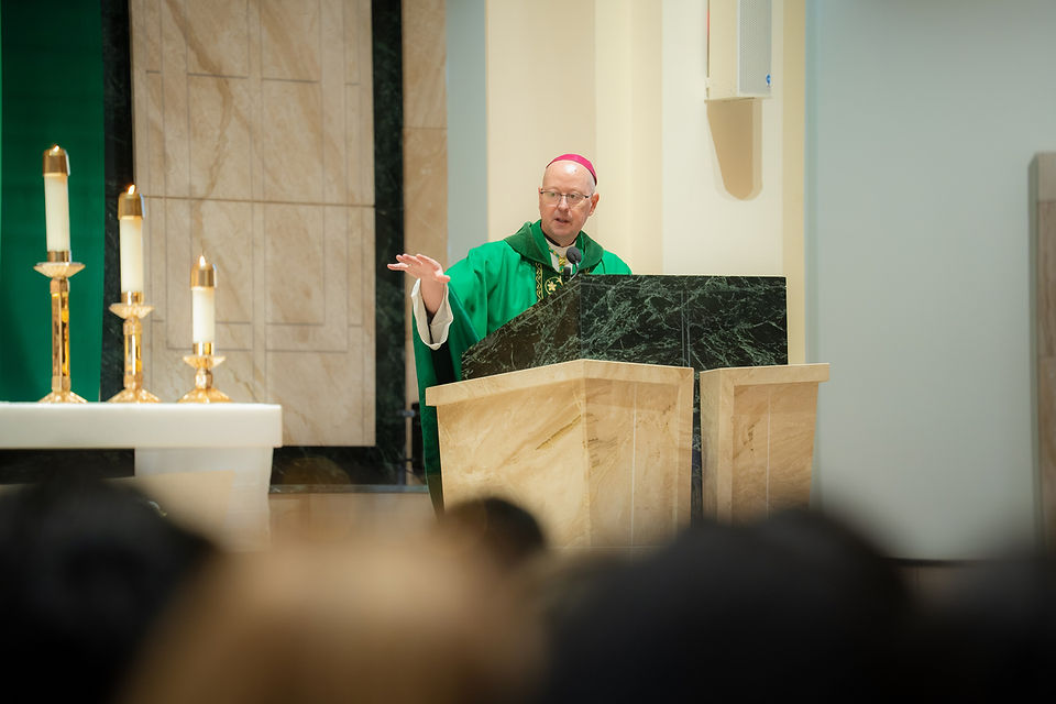 Bishop in green vestments speaks at a church lectern beside lit candles, addressing a quiet congregation.