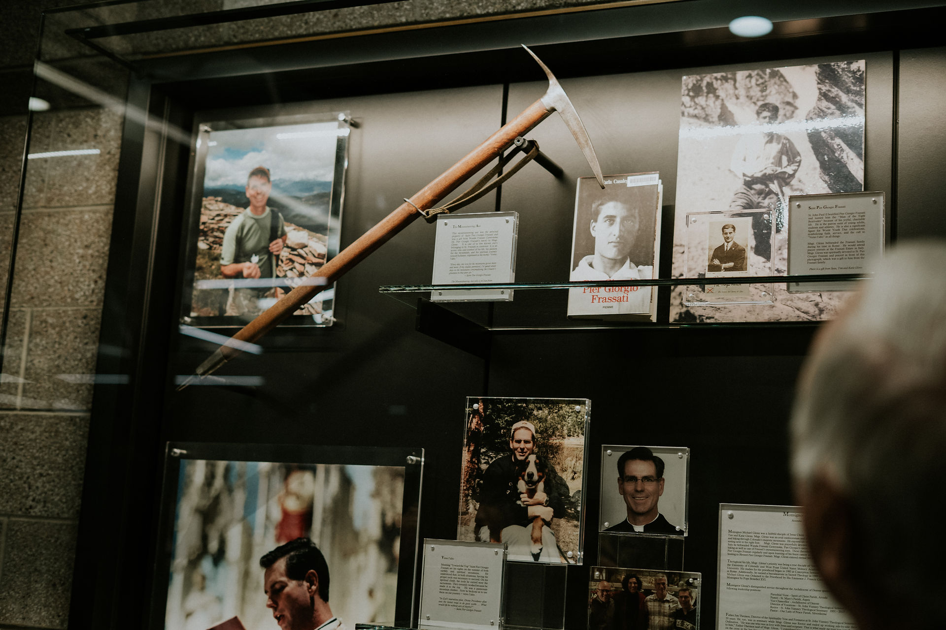 Display case with photos and a vintage ice axe. Text and images of various people, including Pier Giorgio Frassati. Mood: historical.