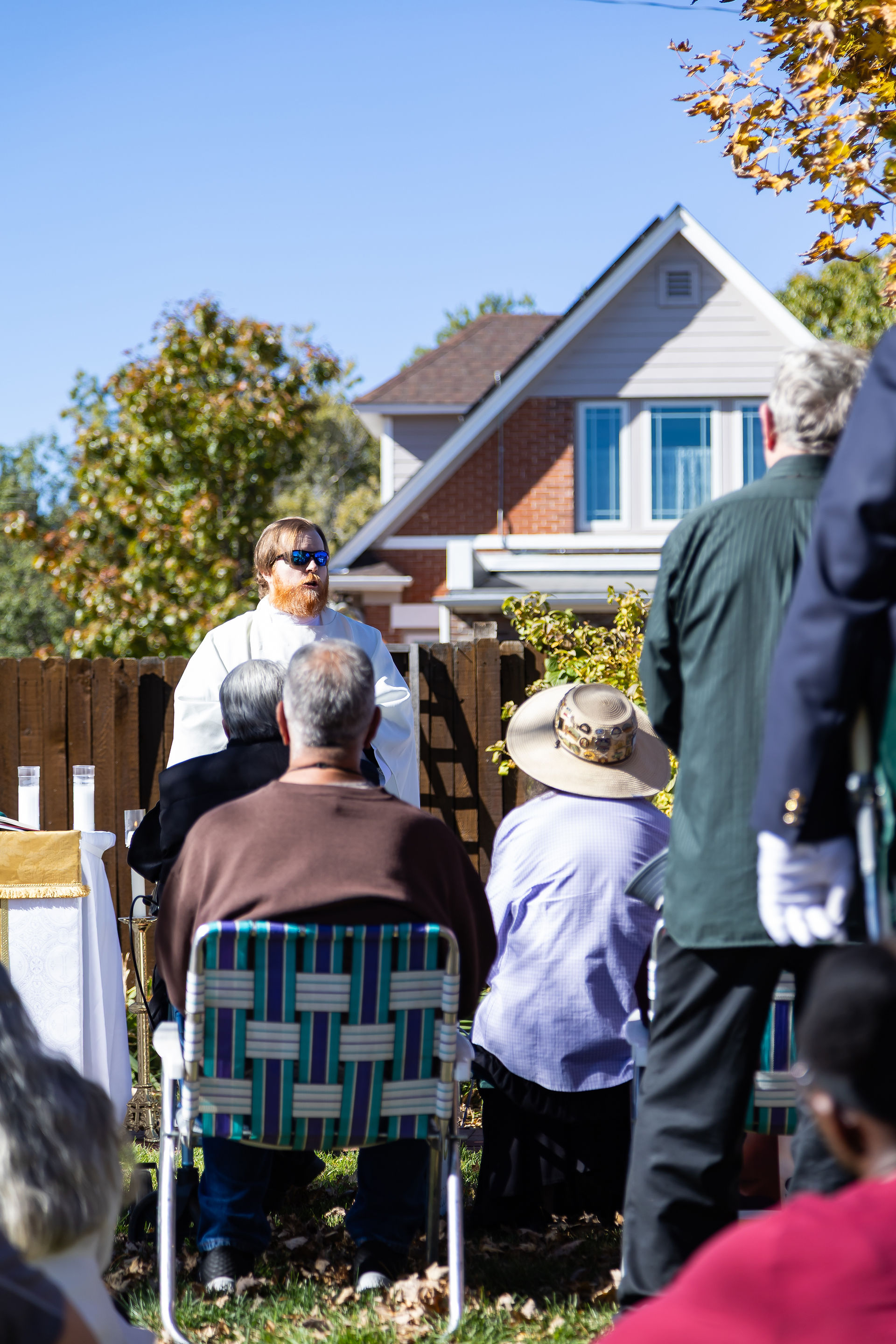 Priest in white robe and sunglasses speaks outside to seated crowd; house and trees in background; sunny, relaxed setting.