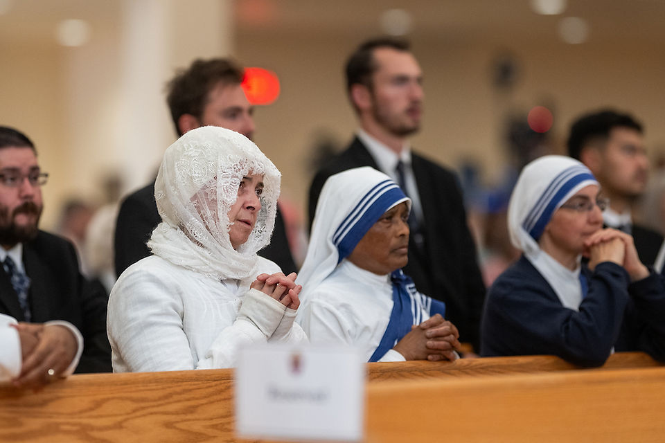 People in religious attire sit in a church, hands clasped in prayer. A "Reserved" sign is visible. The mood is solemn and focused.