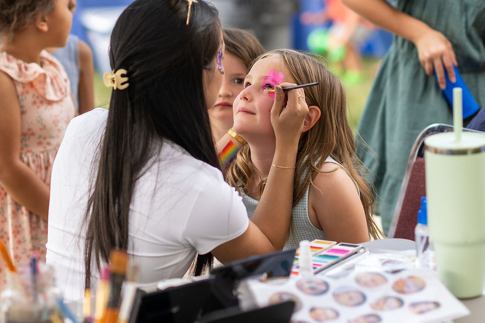 Artist paints a pink star on a young girl’s face at an outdoor fair, with other children watching nearby.