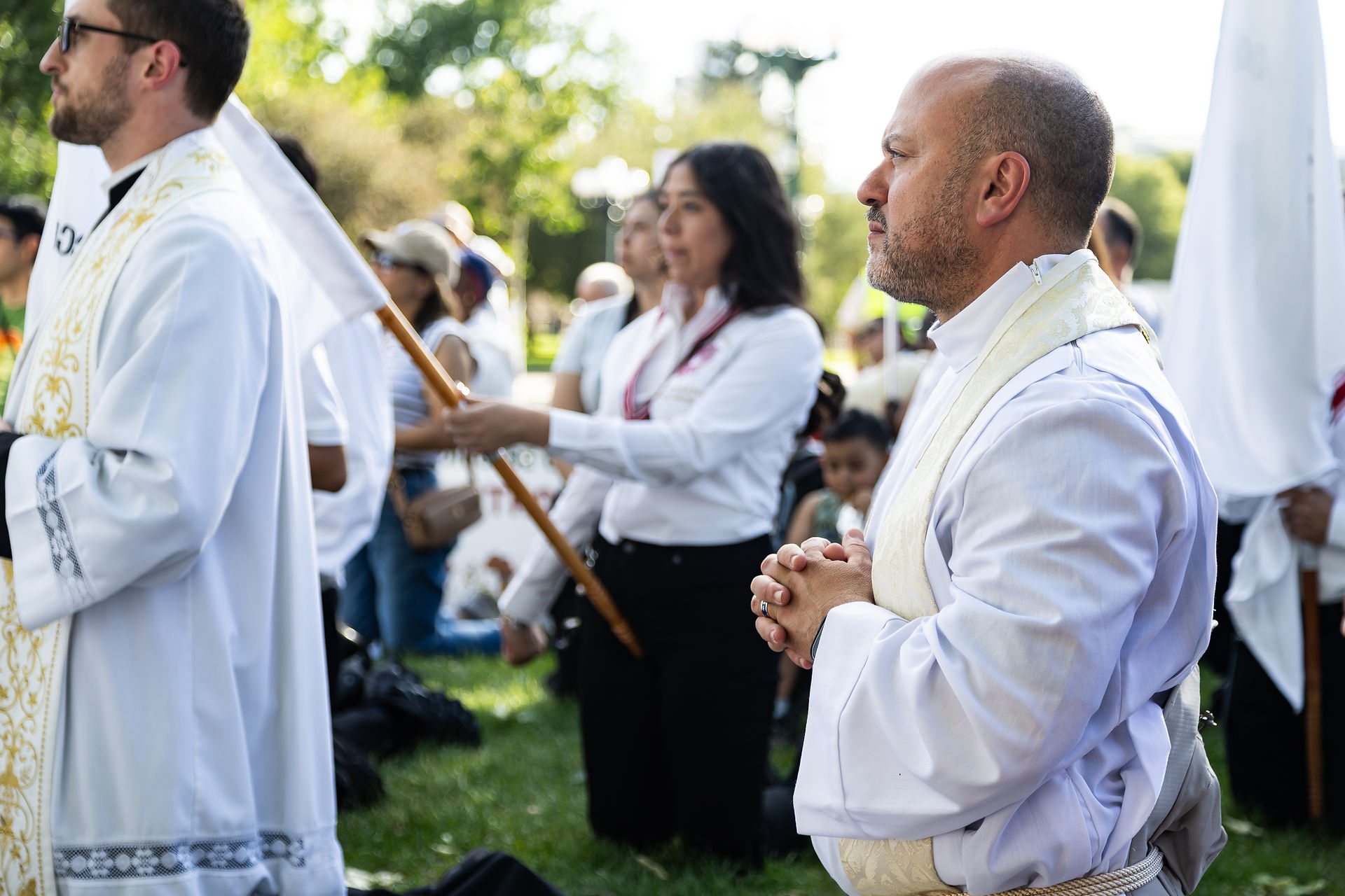 Clergy and worshippers in white robes stand outdoors in prayer, one man clasping his hands during a solemn ceremony.