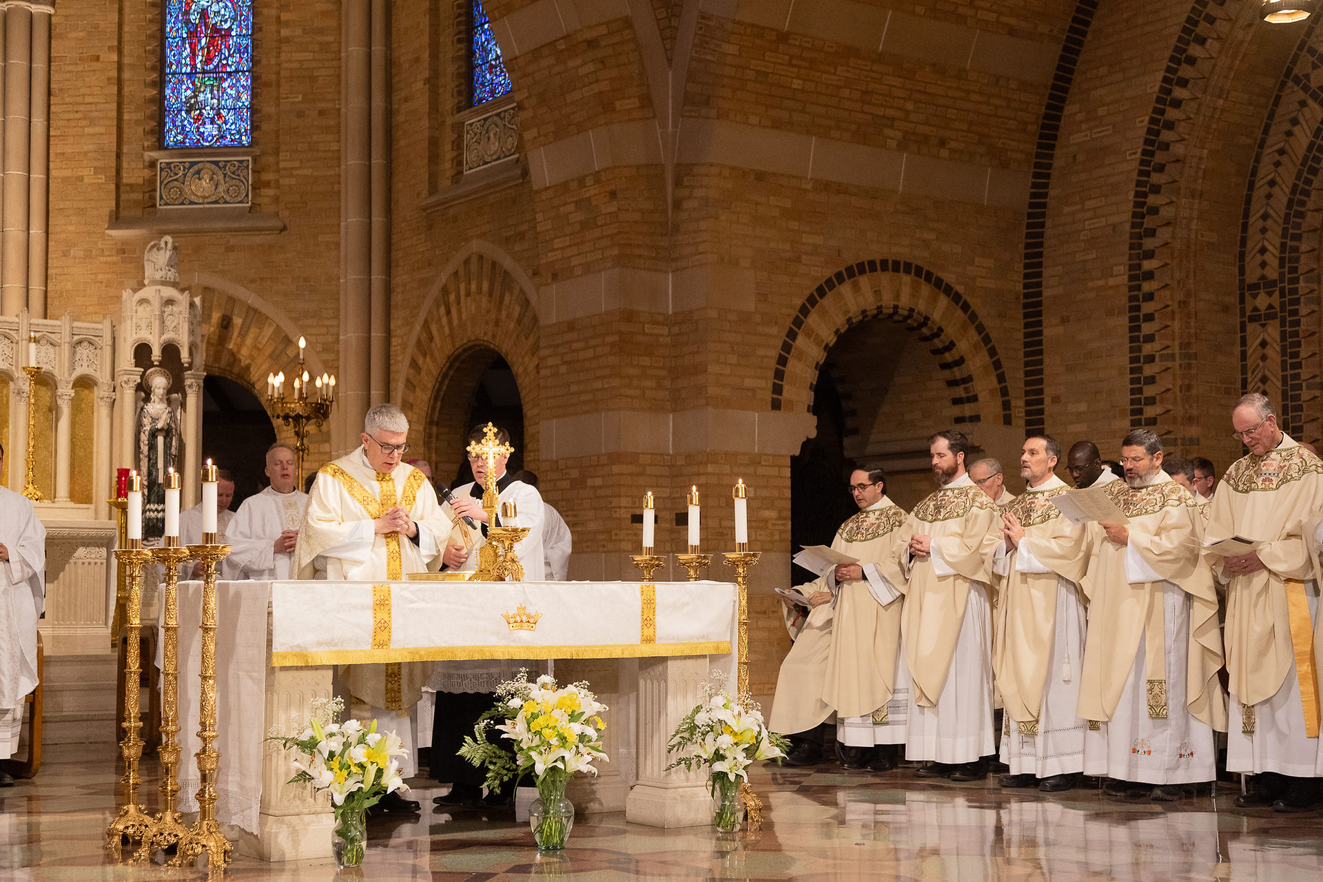 A priest stands at the altar during a religious ceremony, reading from a binder placed upon the altar. Other priests look on in the distance. The background features a beautiful brick chapel and stained glass windows. The mood is celebratory.