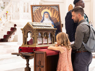 Family prays at a church reliquary beside an icon of a saint, with a man and little girl at the altar.