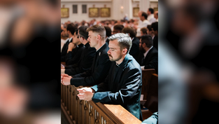 Men in dark suits pray quietly in a sunlit church, standing at wooden pews during a solemn service.