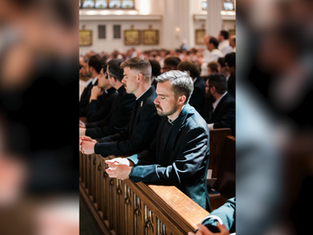 Men in dark suits pray quietly in a sunlit church, standing at wooden pews during a solemn service.
