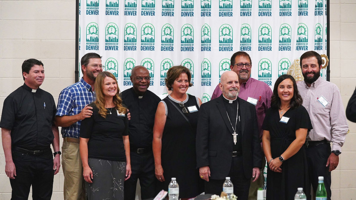 A group of smiling people, including clergy, pose in front of an "Archdiocese of Denver" backdrop in a hall. Bottles on the table.