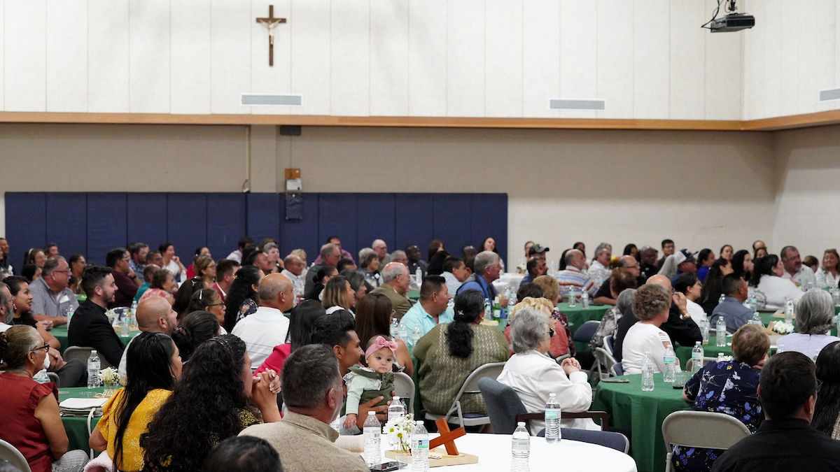 A large group of people seated at round tables with green covers in a hall, listening attentively. A cross decorates the wall above.