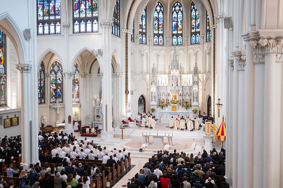 Clergy in white robes gather at an altar in a grand cathedral. Stained glass windows illuminate the ceremony, watched by a large congregation.