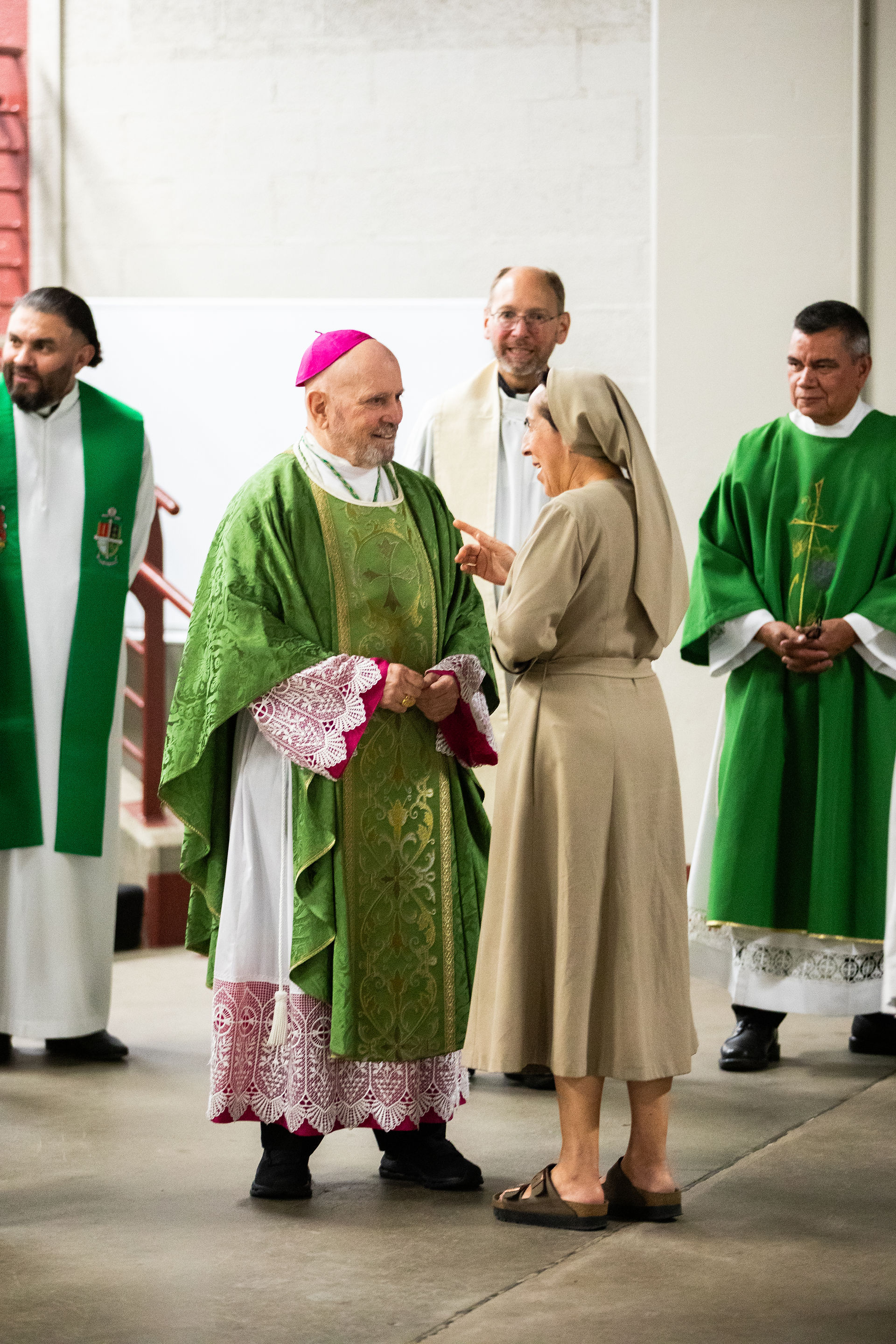 A group of religious figures gathered indoors, including a bishop in a green chasuble with gold embroidery and a magenta zucchetto, a nun in a beige habit and sandals speaking to him, and several priests in green chasubles with white stoles. One priest’s chasuble features a cross. The setting suggests a church or religious building, emphasizing clergy attire and interaction during a religious gathering.